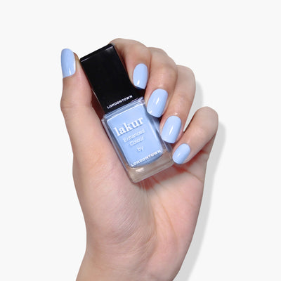 A hand with manicured nails holds a bottle of LONDONTOWN's "In the Clouds" cruelty-free nail polish in light blue.