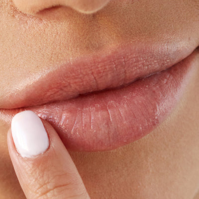 Close-up of a person's slightly parted lips. A finger with white nail polish is gently touching the lower lip, applying LONDONTOWN's Smoothing Lip Balm for an ultra-rich conditioning treatment.