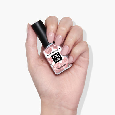 Hand holding a bottle of LONDONTOWN Make Me Blush gel color nail polish with a floral label design. Nails are painted in a light pink shade, showcasing the vibrant, vegan-friendly formula that cures perfectly under UV/LED light.