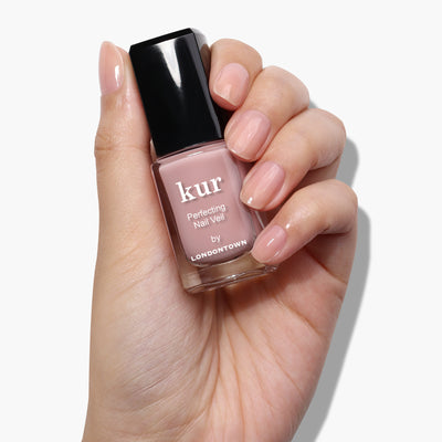 A hand with neatly manicured nails holds a bottle of LONDONTOWN Perfecting Nail Veil #4 nail polish, offering a hint of color in a light pink shade. This power-packed polish not only beautifies but also provides essential care for your nails.