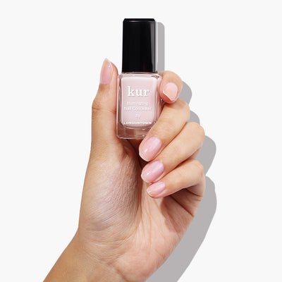 A hand holds a bottle of Pink Illuminating Nail Concealer by LONDONTOWN, its nails boasting a light pink finish that matches the bottle's shade.