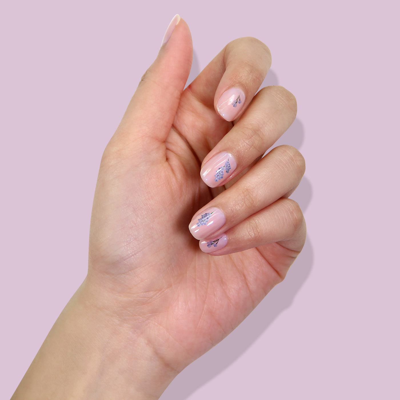 The Nail Decals - Petals in Bloom by LONDONTOWN feature a hand adorned with light pink nail polish and intricate blue floral designs against a light purple backdrop, providing an effortless solution for achieving a chic look.