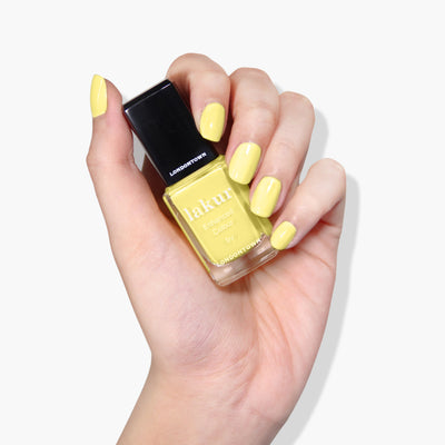 A hand with manicured nails painted in a lemon meringue yellow holds a bottle of cruelty-free LONDONTOWN Lakur nail polish in the shade Notting the Fancy.
