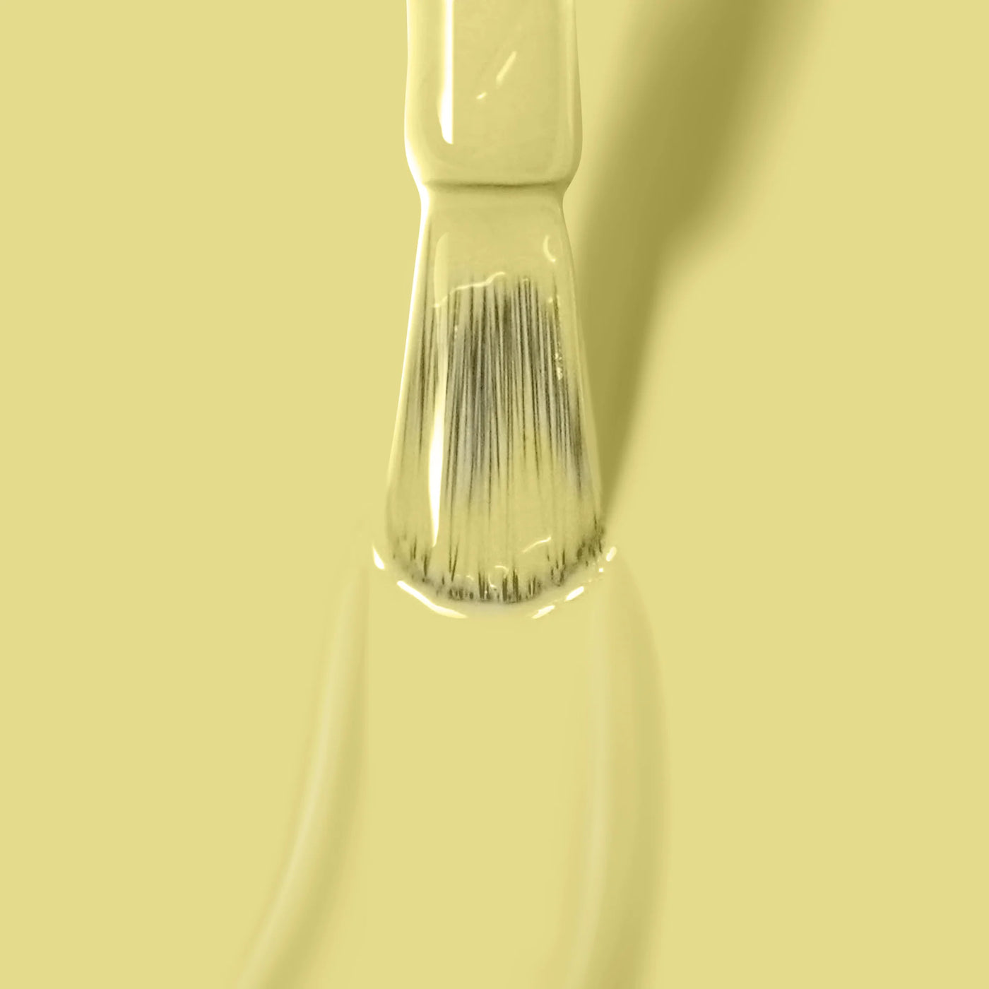 A close-up of a LONDONTOWN Notting the Fancy (Professional) paintbrush with beige bristles, coated in light yellow paint reminiscent of lemon meringue yellow nail polish, set against a matching yellow background.