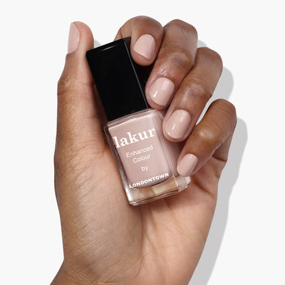 A hand holding a bottle of Pampas Beige by LONDONTOWN nail polish in a creamy beige shade. The nails are painted in the same buff color as the polish.