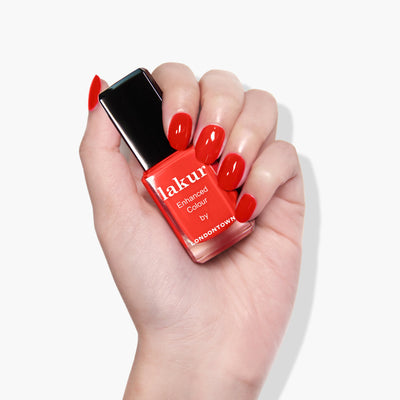 A hand with red nails holds a bottle of ultra-bright blood orange crème nail polish labeled "Piccadilly Square (Professional) by LONDONTOWN." This vegan, cruelty-free nail polish is part of the luxurious Londontown polishes collection.