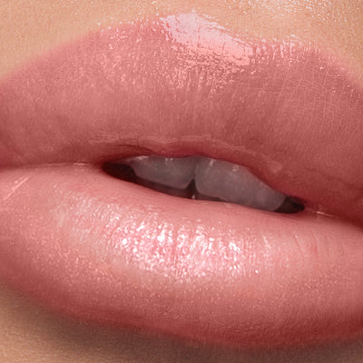 A close-up image shows glossy, slightly parted milky pink lips with visible texture and a hint of teeth, enhanced by LONDONTOWN's Plush Shine Lip Gloss in Pixie.