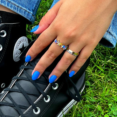 A hand with nails painted in "Poolside Dreams (Professional)," adorned with two rings, rests on a pair of black Converse sneakers. The person is sitting on grass, showing off their vibrant *vegan nail polish* from LONDONTOWN.