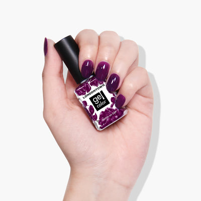A hand with purple nail polish holds a LONDONTOWN gel color bottle in an elegant shade named Portobello Plum, a deep warm plum crème.