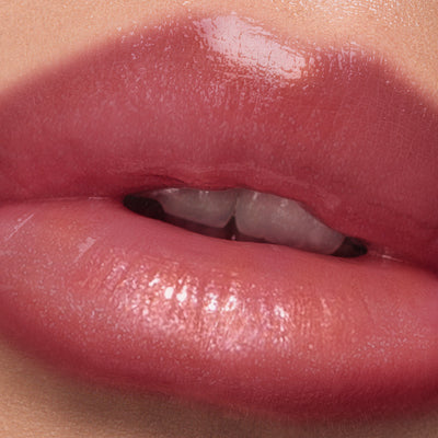 A close-up of glossy, flushed pink-tinted lips enhanced by LONDONTOWN's Plush Shine Lip Gloss in Posh, with slightly parted teeth visible.