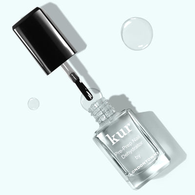 A bottle of LONDONTOWN Pre-Prep Nail Dehydrator with its brush applicator unscrewed, lying on a light green surface with a few clear droplets nearby, sets the stage for next-level professional polish prep.