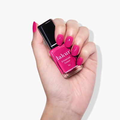 A hand with long, manicured nails is holding a bottle of pink nail polish labeled "Queen of Hearts by LONDONTOWN." The vegan, cruelty-free polish is a deep fuchsia pink crème, and the nails are painted in the same vibrant shade.