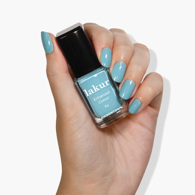 A hand with teal-colored painted nails is holding a bottle of LONDONTOWN's Lakur vegan nail polish in the shade Reverse the Charges.
