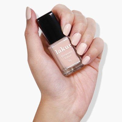 A hand with manicured nails painted in a nude shade holds a bottle of Sandstone nail polish by LONDONTOWN, featuring the nourishing Florium Complex.