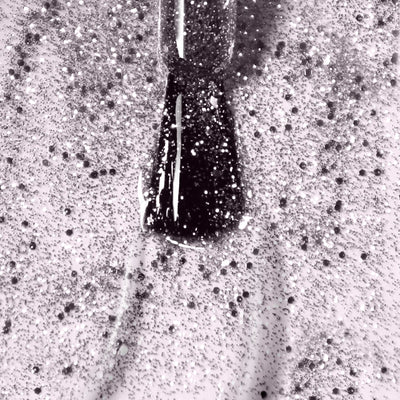 A close-up image of a nail polish brush applying LONDONTOWN's Starstruck (Professional), a vegan, cruelty-free silver glitter nail polish, on a surface. The shimmer polish’s glitter particles are scattered across the thin layer of polish.