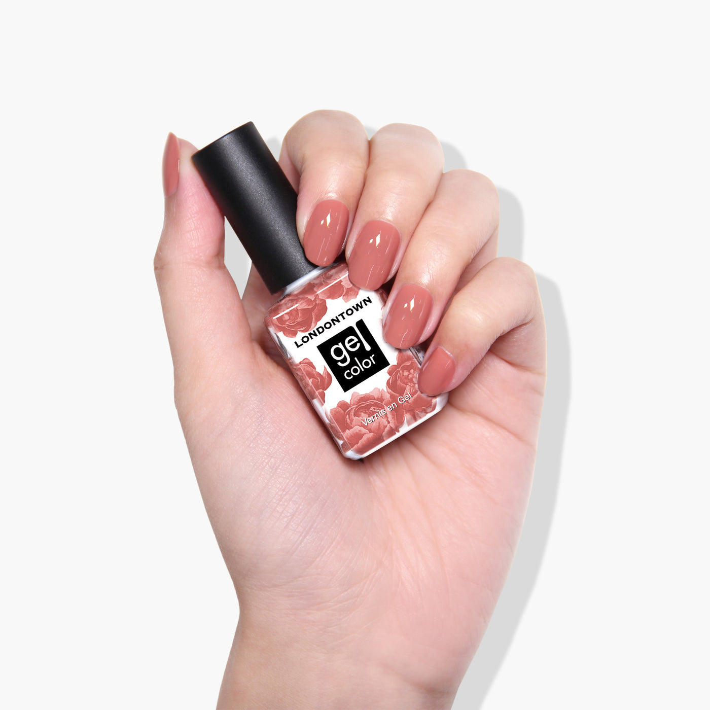 A hand holding a bottle of LONDONTOWN's Toffee Tart gel nail polish in a pinkish-nude shade with a black cap. The nails, painted in the same Toffee Tart gel color, shimmer subtly under UV/LED light.