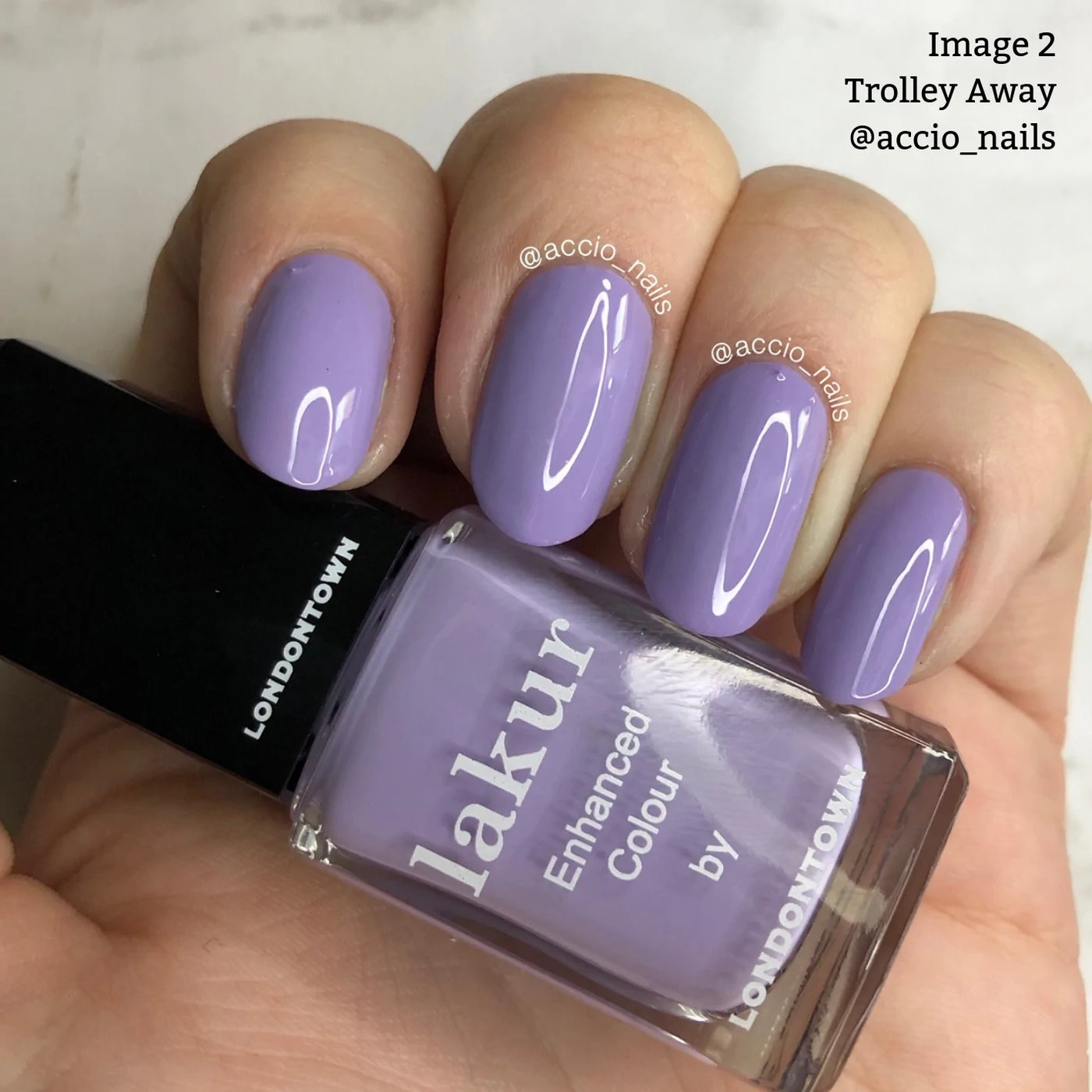 Close-up of a hand with manicured nails painted in a lilac crème nail polish shade called "Trolley Away (Professional)" by LONDONTOWN. This cruelty-free and vegan polish is held in the image. Text reads, "Image 2 Trolley Away @accio_nails.