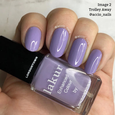 Close-up of a hand with manicured nails painted in a lilac crème nail polish shade called "Trolley Away (Professional)" by LONDONTOWN. This cruelty-free and vegan polish is held in the image. Text reads, "Image 2 Trolley Away @accio_nails.