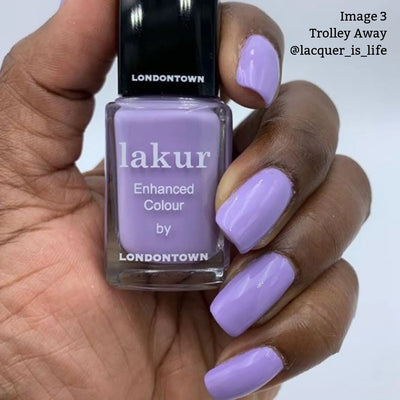 A hand with manicured nails painted in a lilac crème shade holds a bottle of Londontown Trolley Away (Professional) nail polish in the same color. Text on the bottle reads, "lakur Enhanced Colour by LONDONTOWN." The polish is both cruelty-free and vegan.