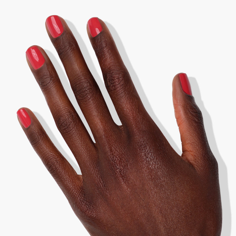 A sun-kissed hand with a dark skin tone and neatly manicured nails painted in LONDONTOWN's Flushed Cheeks, a vibrant red shade, set against a white background.