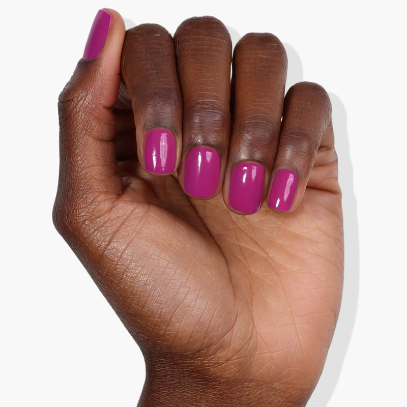 A hand with dark skin tones displays fingernails painted in a glossy shade of Violet Hibiscus, part of LONDONTOWN's collection, beautifully accentuated by deep ultraviolet hues against a white background.