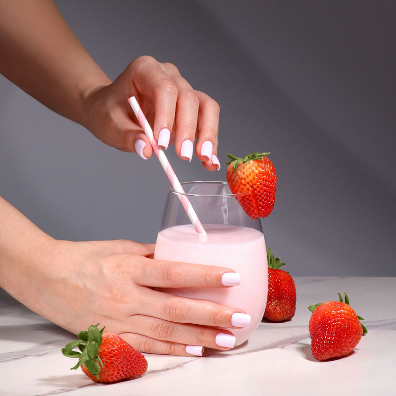 A person with manicured nails holds a glass of LONDONTOWN Strawberry Milk with a striped straw, surrounded by fresh strawberries on a white surface.