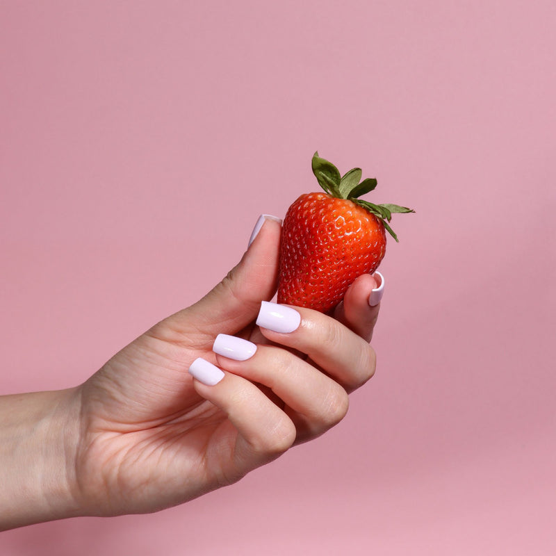 A hand with LONDONTOWN's Strawberry Milk nail polish, a milky light pink shade, holds a sweet red strawberry against a solid pink background.