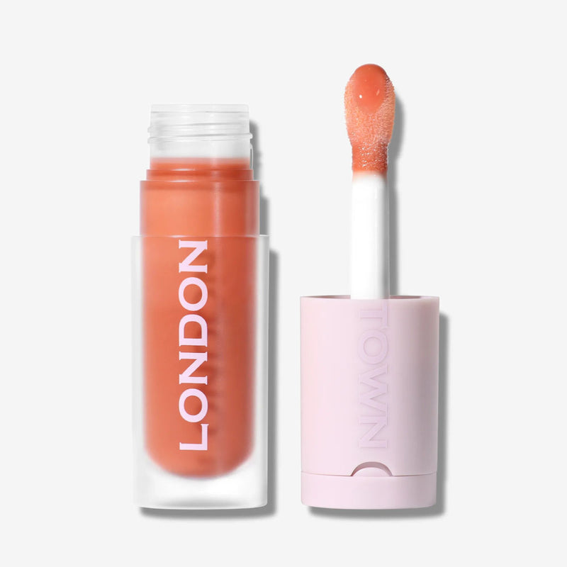 A tube of LONDONTOWN Quench Glow Buttery Lip Oil in Honeyed, with "LONDON" written vertically on the side and the applicator upright beside it, delivers plush shine and a sheer peachy tint to your lips.