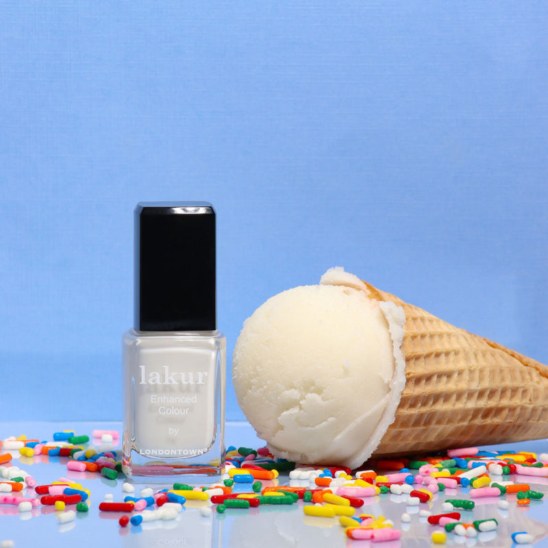 A bottle of LONDONTOWN’s Double Scoop creamy white nail polish stands beside a vanilla ice cream cone with colorful sprinkles, set against a blue background.