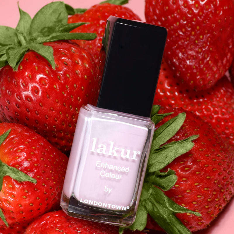 A bottle of LONDONTOWN Strawberry Milk nail polish rests atop a pile of fresh strawberries.