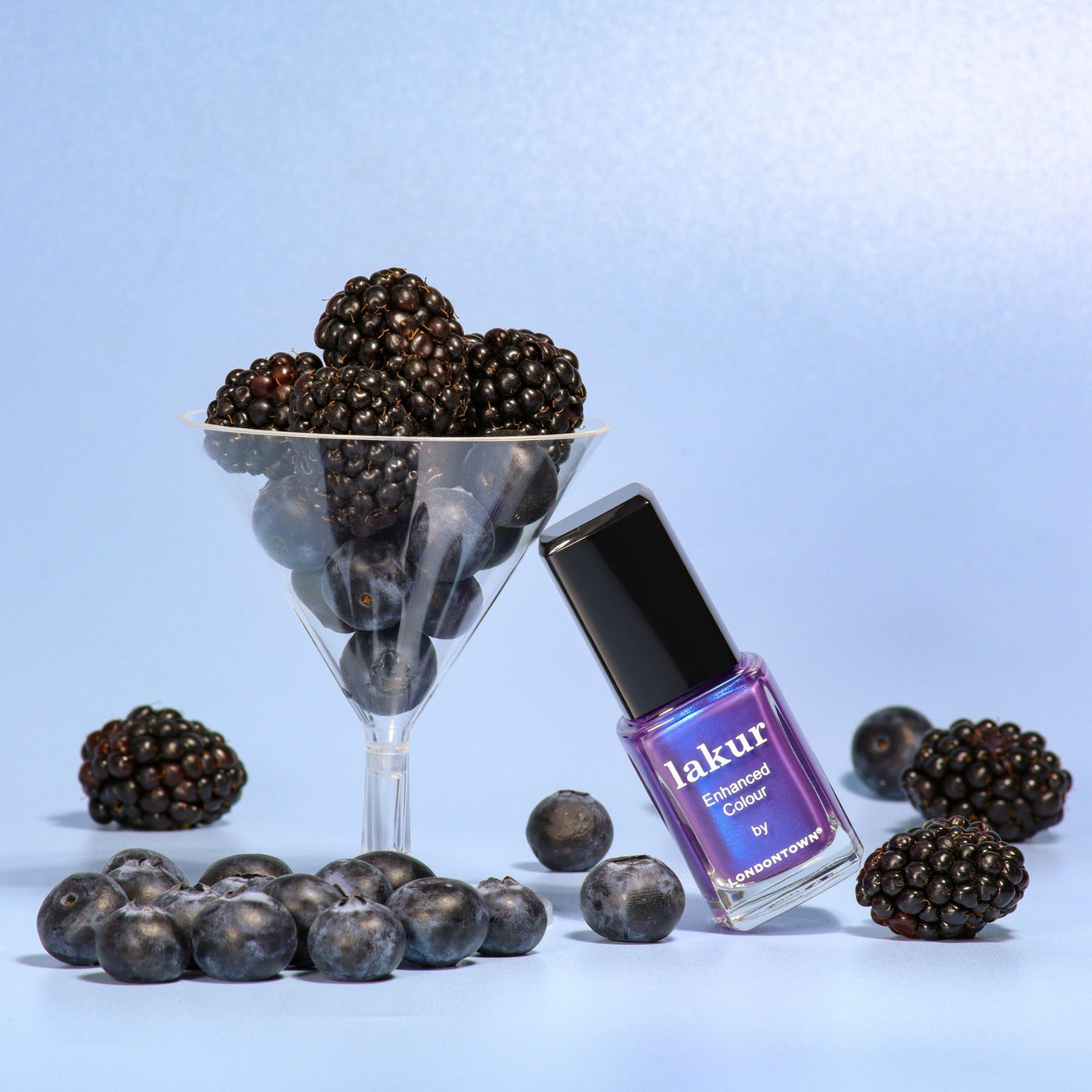 A bottle of LONDONTOWN Wildberry Freeze nail polish with blue shimmer sits on a blue background, next to a martini glass filled with blackberries and scattered blueberries.