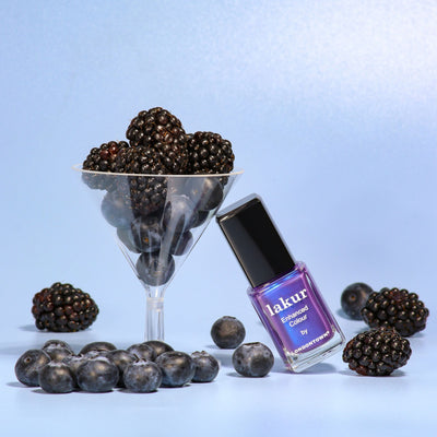 A bottle of LONDONTOWN Wildberry Freeze nail polish with blue shimmer sits on a blue background, next to a martini glass filled with blackberries and scattered blueberries.