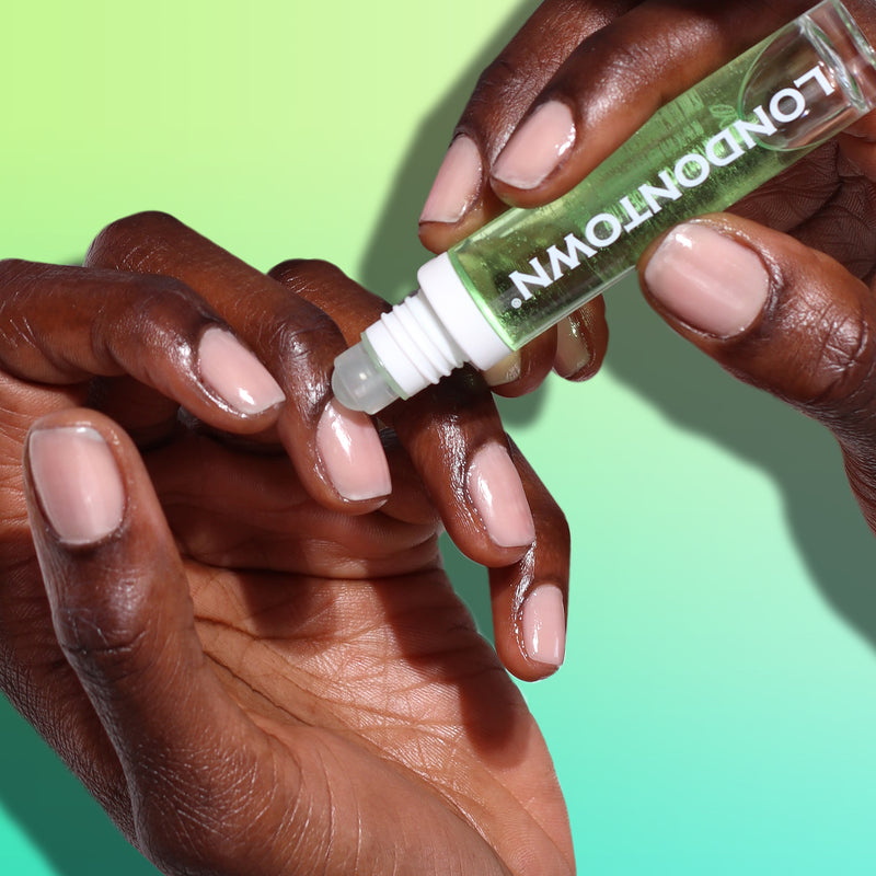 Hands applying the Roll & Glow Cuticle Oil - Agave Pear from a pocket-sized roll-on bottle labeled "LONDONTOWN" to manicured nails, against a gradient green background.