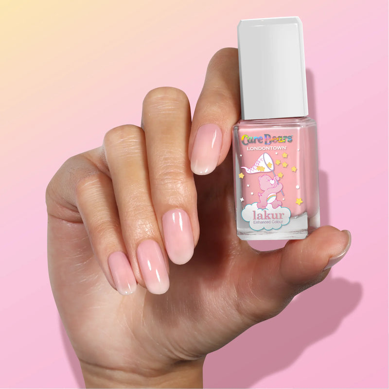 A hand with natural, glossy nails holds a bottle of LONDONTOWN "BFF" sheer pink nail polish against a pink gradient background.