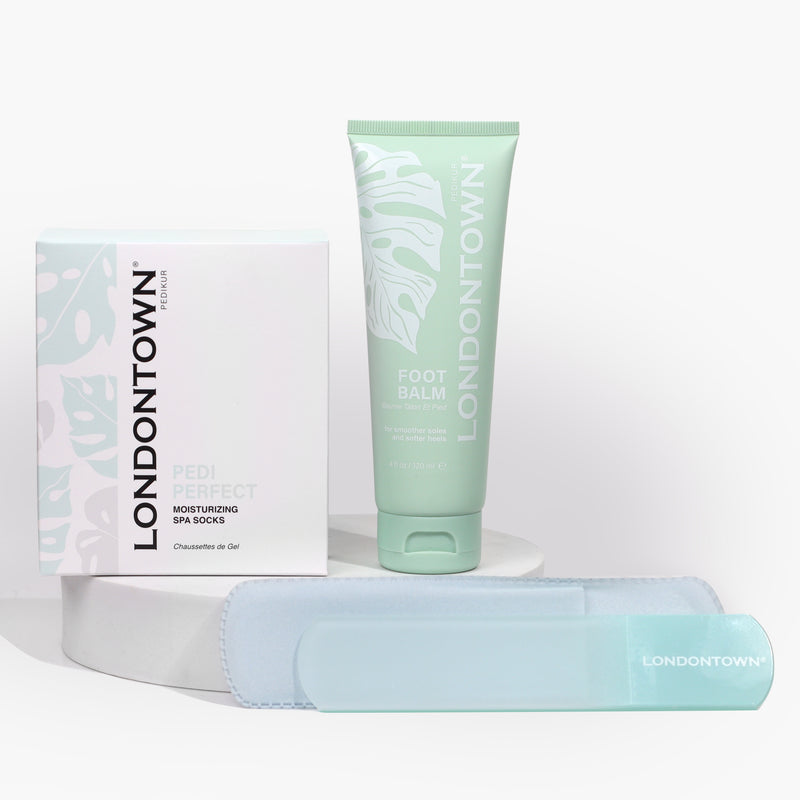 The LONDONTOWN Sole Soothing Trio, designed for a complete pedi refresh, includes moisturizing spa socks, foot balm, and a foot file. All items come in chic mint green packaging and are elegantly displayed on a white platform.