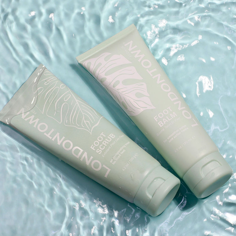 Two mint green LONDONTOWN Pedi Discovery Sets, featuring a foot scrub and a foot balm, lay side by side on a rippling aqua background.