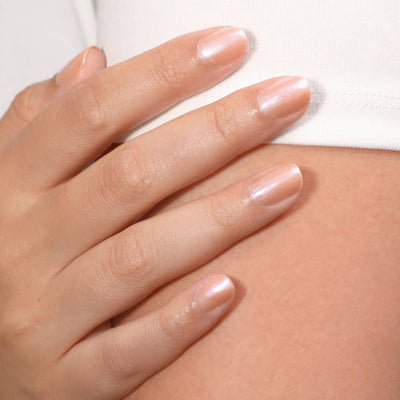 A close-up of a hand with neatly manicured nails, showcasing the cult-favorite Bare Illuminating Nail Concealer (Professional) by LONDONTOWN in a lustrous beige shade, resting on light-colored fabric.