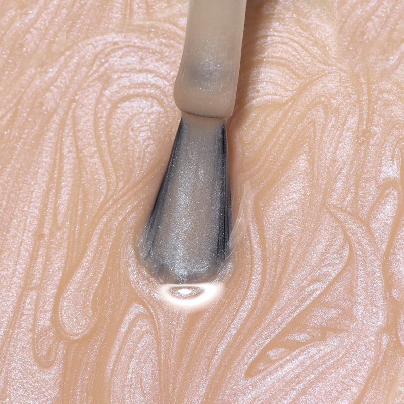 A close-up of a LONDONTOWN Bare Illuminating Nail Concealer brush applying pearlescent nude polish on a textured surface.