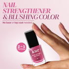 A hand with manicured nails holds a bottle of LONDONTOWN Sheer Strength Nail Blush - Hibiscus, a 2-in-1 nail hardener and blushing color that strengthens nails while adding a glow-boosting pink tint.