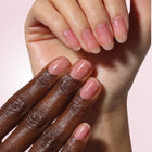 Two hands, one dark-skinned and one light-skinned, display neatly manicured nails against a soft pink background—highlighting the results of LONDONTOWN Sheer Strength Nail Blush in Hibiscus.