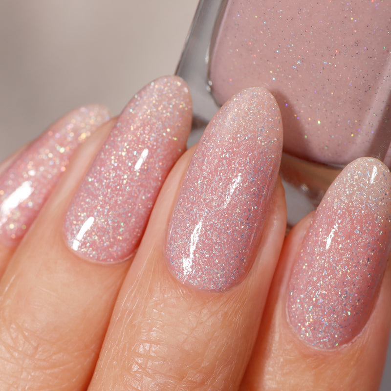 A hand with holographic glittery pink nails, wearing Champagne in Bed by LONDONTOWN, elegantly holds the polish bottle.