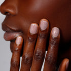 A close-up shows a person touching their face, showcasing manicured short nails in "Champagne in Bed," a neutral pink with subtle sparkle by LONDONTOWN, against dark skin.