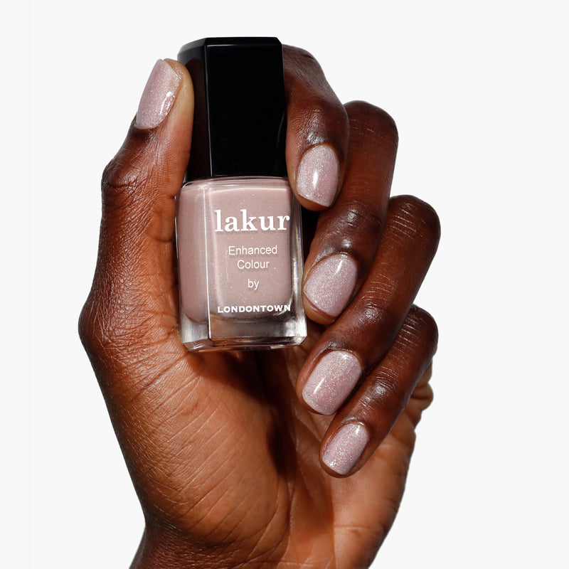 A hand holds a bottle of LONDONTOWN's Lakur nail polish in the shade Champagne in Bed, with nails painted to match this neutral pink hue that offers a subtle holographic sparkle.