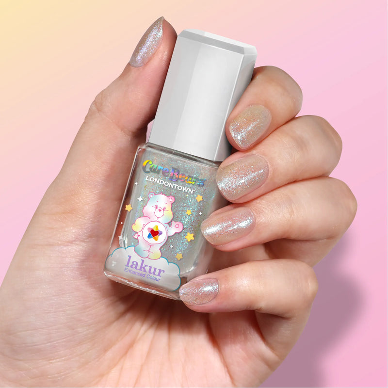 A hand with glittery nail polish holds a bottle of LONDONTOWN’s Chasing Rainbows, its iridescent shimmer and Care Bears design evoking a sense of hope against a soft pink backdrop.
