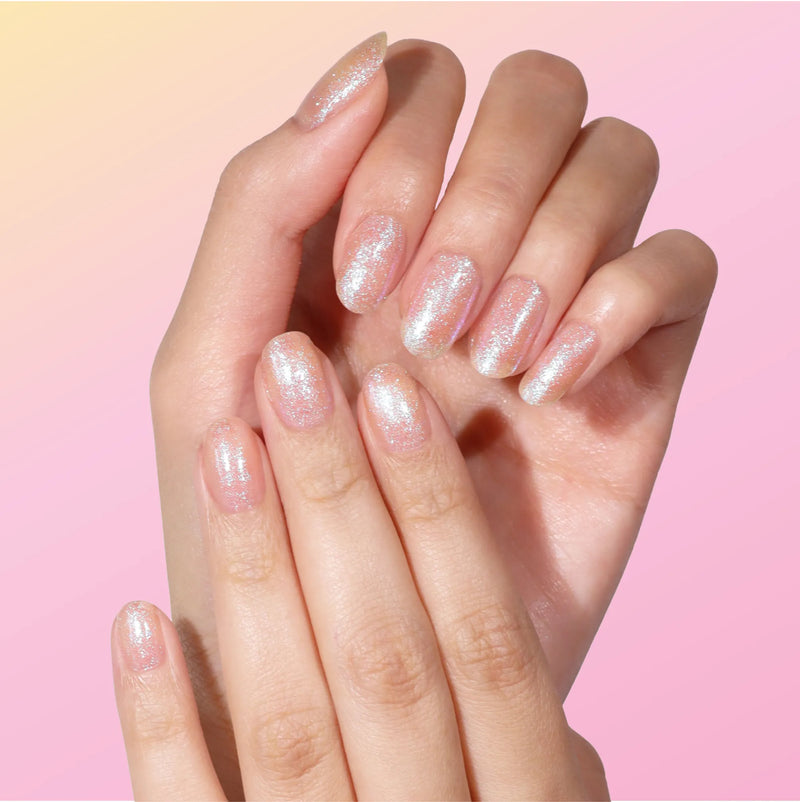 A person shows off neatly manicured, long oval nails painted with LONDONTOWN's Chasing Rainbows in an iridescent shimmer, set against a pink and yellow gradient background that suggests soft hopefulness.
