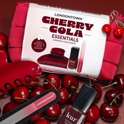 LONDONTOWN’s Cherry Cola Essentials set includes nail polish, nail file, hair clip, and cherries, capturing the deep tones of Cola Crush against a vivid red backdrop.