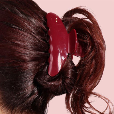 A section of dark red hair, styled with the LONDONTOWN Cherry Cola Essentials set, is held in a large maroon claw clip against a light pink background.