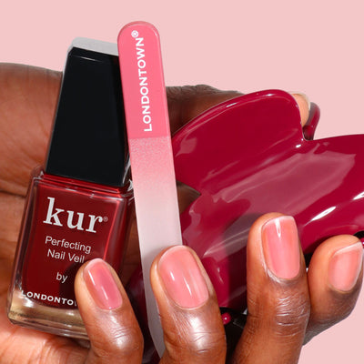 A hand holds LONDONTOWN’s Cherry Cola Essentials—a bundle featuring polish, a red "LONDONTOWN" nail file, and a red polish spill—against a pink background.