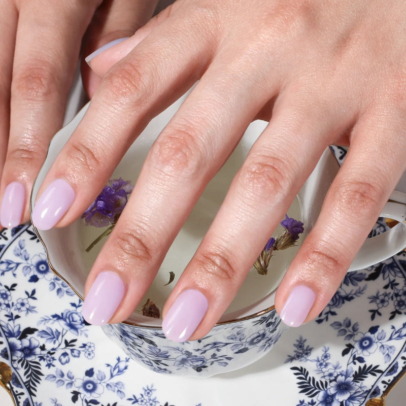 A person with Cloud Nine by LONDONTOWN periwinkle nails rests their hand on a blue and white patterned teacup and saucer filled with small purple flowers.