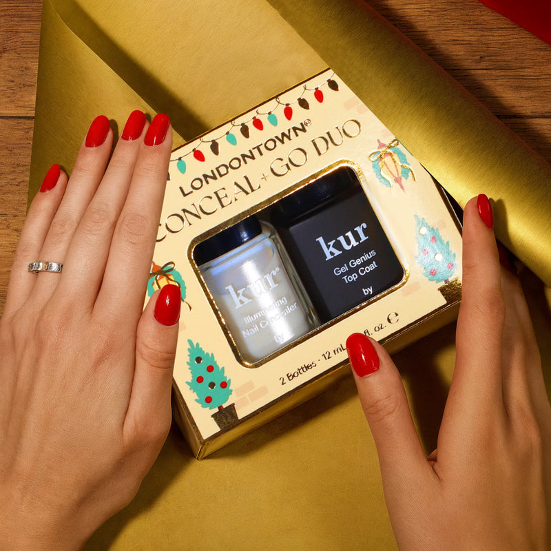 Hands with red nail polish hold LONDONTOWN’s Conceal + Go: Holiday Edition nail care set—your secret to naturally perfect nails—against a wooden surface and gold wrapping paper.